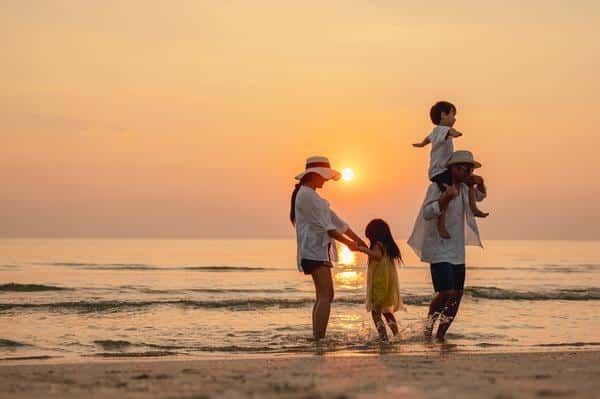 Family of four enjoying a sunset at the beach: two parents in shallow water with their kids, one child on a parent's shoulders in the orange glow.