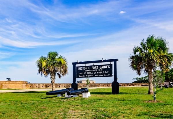 Historic Fort Gaines sign on a grassy lawn with two cannons and palm trees under a blue sky, a coastal fort setting.