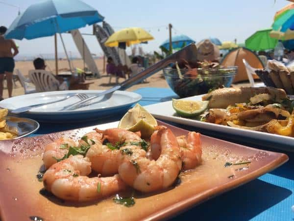 Plate of shrimp with herbs and lime on a sandy beachside table, colorful umbrellas in the background.