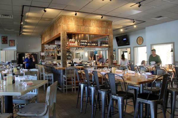 Casual restaurant interior with a wooden central bar, metal chairs, and customers seated at tables.