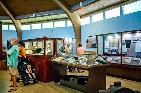 Museum gallery with glass display cases and framed photos on the walls; visitors, including a masked man and a child in a stroller, examining the exhibits.