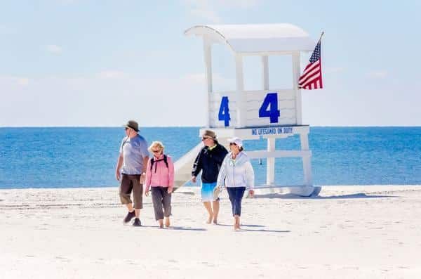 Four adults walking along a sunny beach near a lifeguard tower with an American flag.